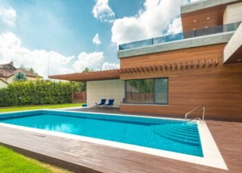 Backyard swimming pool with clear blue water beside a home under blue sky