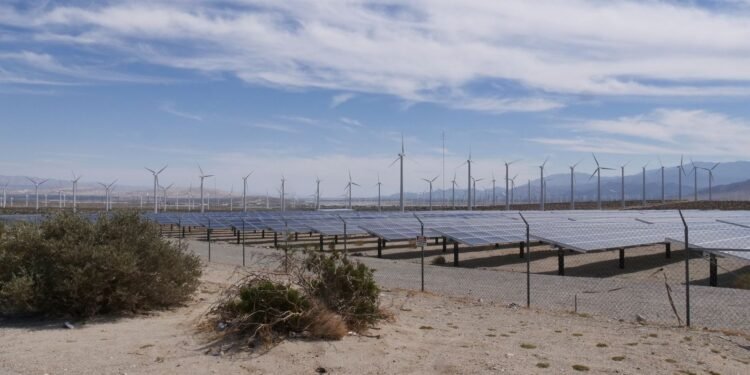 Ground-mounted solar panels with wind turbines across open flat countryside under a wide sky